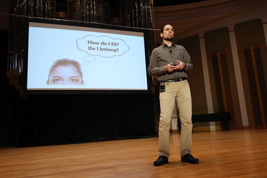 Paul Dean presenting on stage in front of a screen containing the text "How do I fit? Do I belong?"